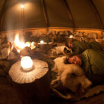 People sleeping on the reindeer skins inside a traditional Sami lavvo tent with the burning fireplace in the middle.