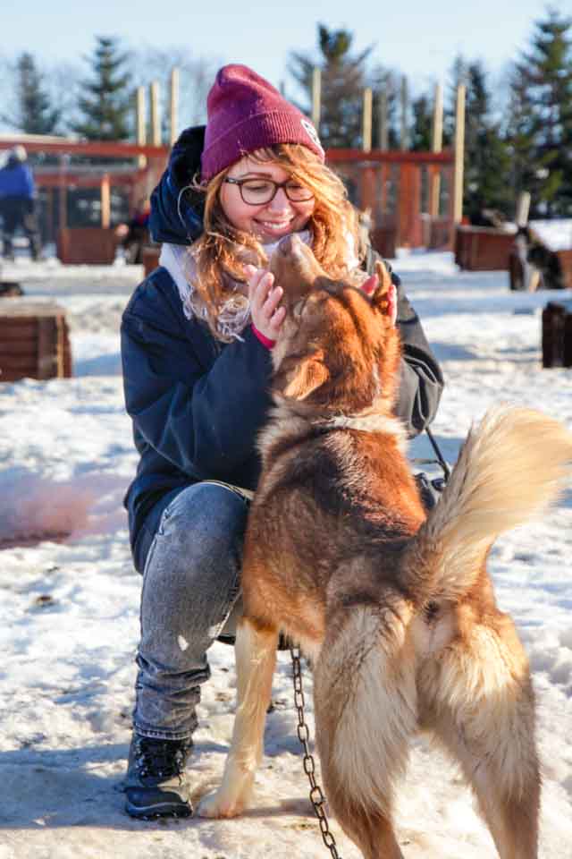 A smiling woman wearing glasses and winter clothes cuddles a ginger dog, gently holding its face. The snowy winter environment adds a cozy and joyful atmosphere to the scene.